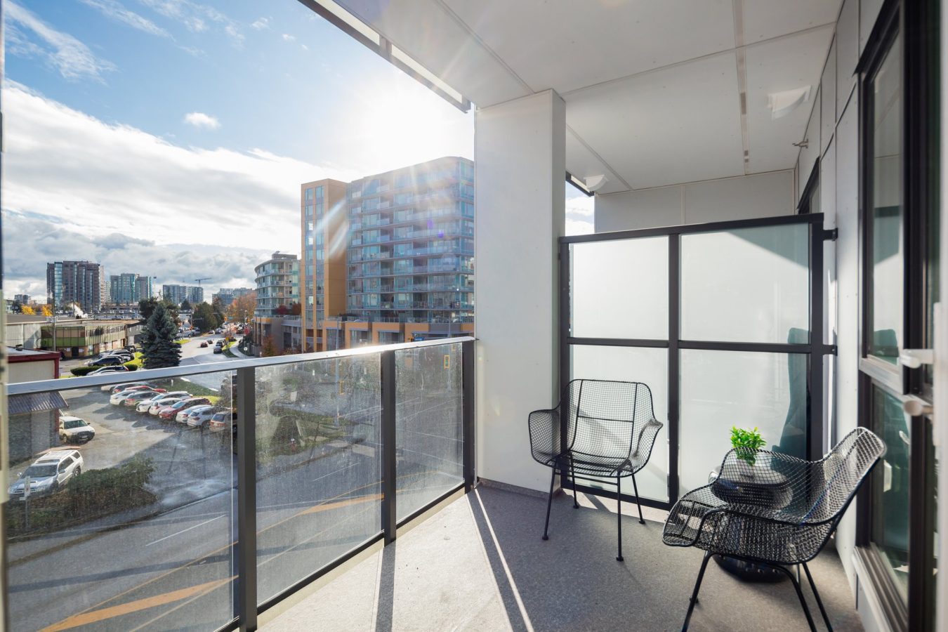 A modern apartment balcony with two black chairs, a small table with a plant, glass railing, and city buildings in the background on a sunny day.