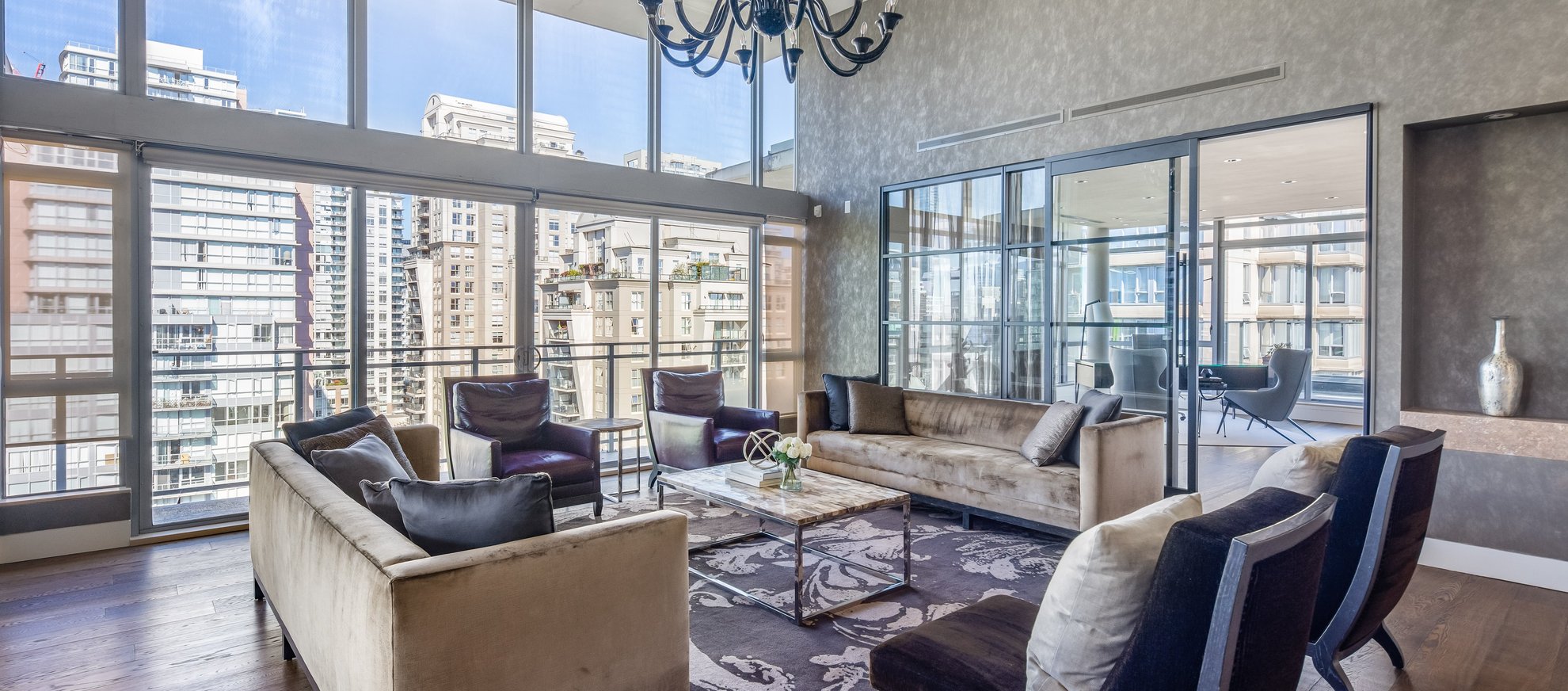A modern living room with large floor-to-ceiling windows, city views, neutral sofas, armchairs, a patterned rug, marble coffee table, and a chandelier. Glass walls divide the living area from an adjacent room.