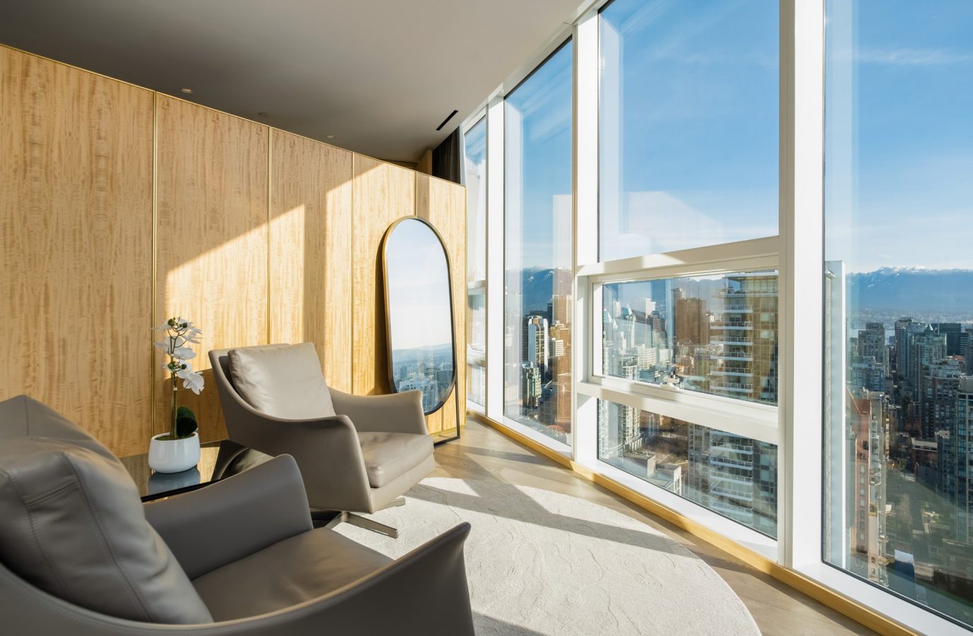 A modern lounge area with two gray armchairs, a round floor mirror, a small table with a potted plant, wood-paneled walls, and large windows offering a city view with mountains in the distance.