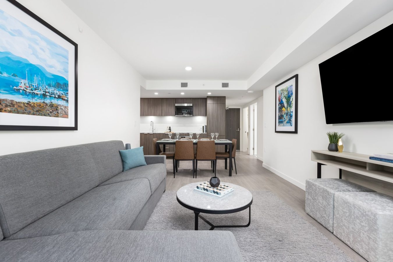 Modern living room with a gray sofa, round coffee table, wall-mounted TV, and dining area in the background. Bright, minimalist décor with framed art and an open kitchen visible.