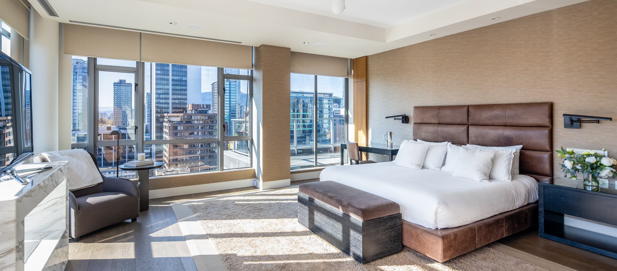 Modern bedroom with large windows showing a city view, a brown upholstered bed with white bedding, a padded bench at the foot, side tables, an armchair by a marble-topped desk, and neutral-colored decor.