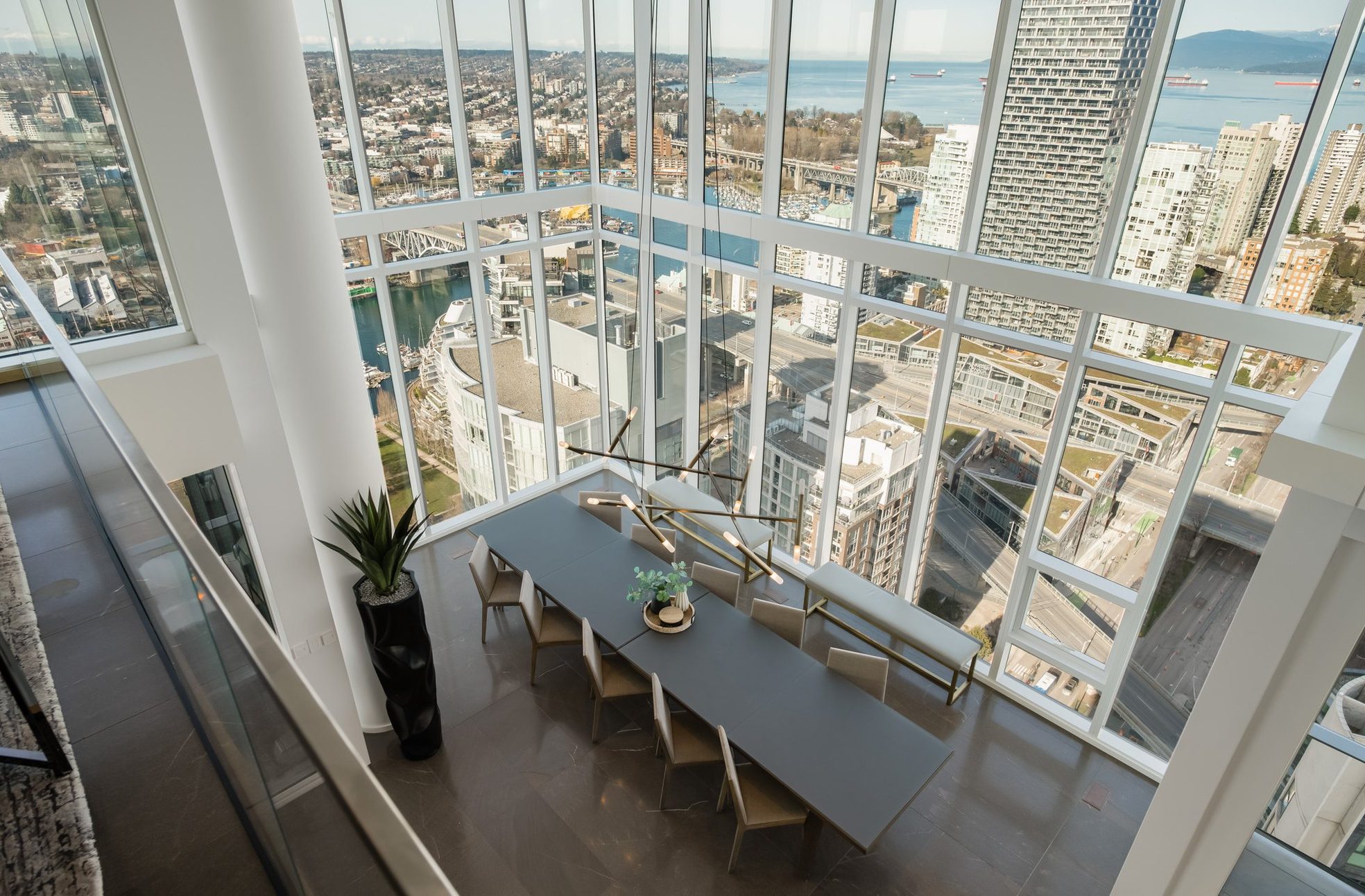 Modern dining area with a long table and chairs, surrounded by floor-to-ceiling windows offering panoramic city and waterfront views from a high-rise building. A plant decorates one corner of the room.