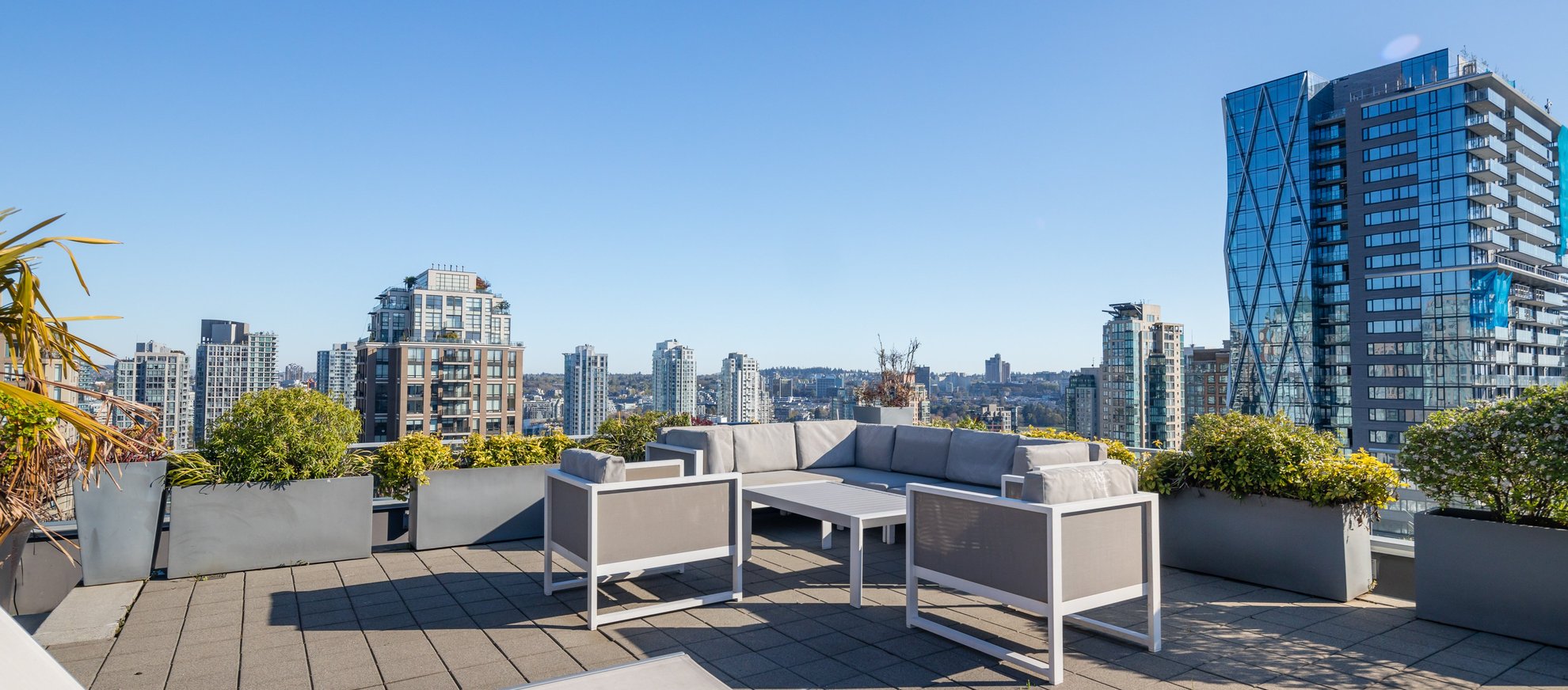 Rooftop patio with modern gray and white outdoor furniture, surrounded by planters, overlooking a city skyline with multiple high-rise buildings under a clear blue sky.