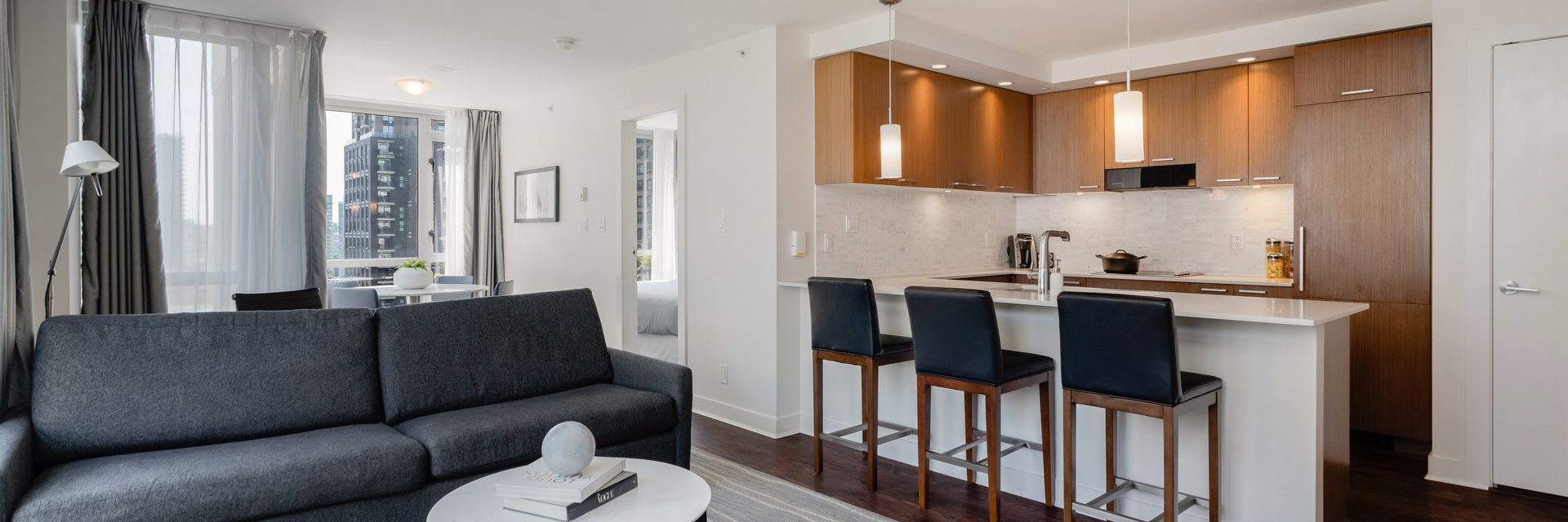 Modern open-concept apartment with a kitchen featuring wooden cabinets, island with three barstools, and pendant lights. Living area has a gray sofa, coffee table, and large windows letting in natural light.