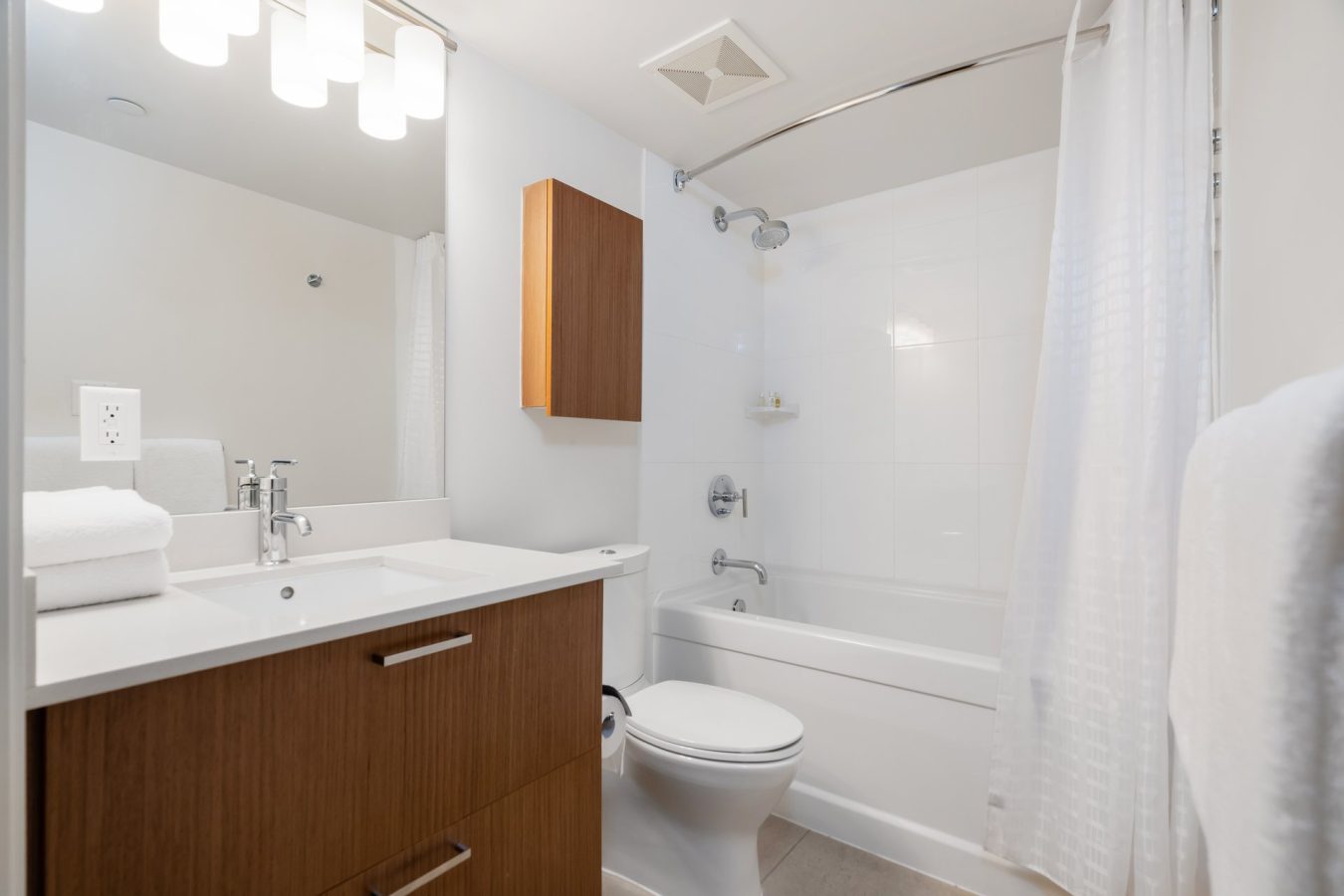 Modern bathroom with a white bathtub and shower curtain, wooden vanity with sink and mirror, wall-mounted cabinet, toilet, towels, and bright overhead lighting. Surfaces are clean and minimalistic.