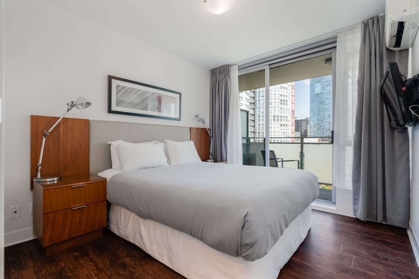 Modern bedroom with a large bed, gray bedding, two wooden nightstands with lamps, wall art above the headboard, a wall-mounted TV, and a sliding glass door leading to a balcony with city views.