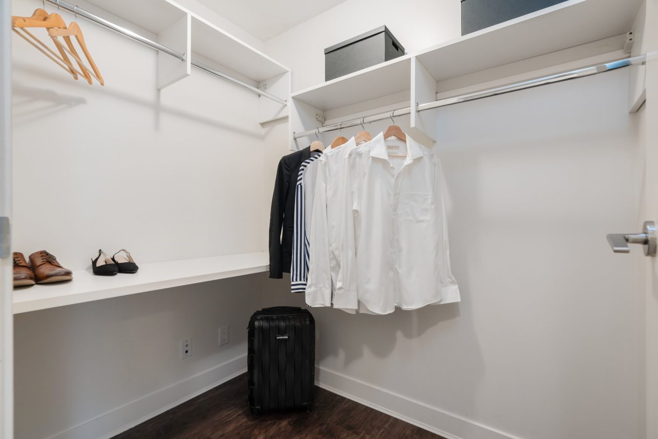 A neatly organized walk-in closet with white shelves and hanging rods, several shirts and a striped sweater on hangers, two pairs of shoes, black storage boxes, and a black suitcase on a dark wood floor.