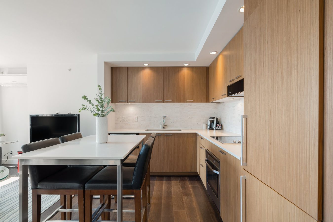 Modern kitchen with light wood cabinets, white countertops, built-in appliances, a center island with black chairs, and a vase with greenery on the table. The space is bright and open with ample lighting.