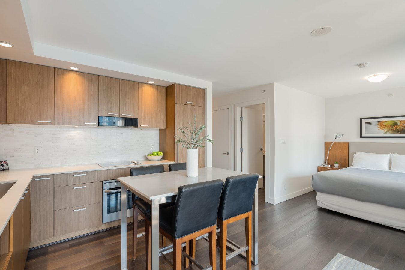 Modern studio apartment with a wooden kitchen, island dining area with three black chairs, and a bed in the background. Room is well-lit with white walls, dark flooring, and minimalist decor.