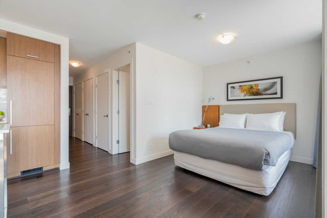 Modern bedroom with dark wood floors, a double bed with white and gray bedding, a bedside table with a lamp, minimalist decor, white walls, and a hallway with several closed doors.