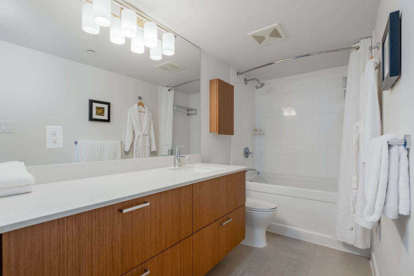 Modern bathroom with a large mirror, wooden vanity, white countertop, bathtub with shower, white towels, a bathrobe hanging on the wall, and bright overhead lighting.