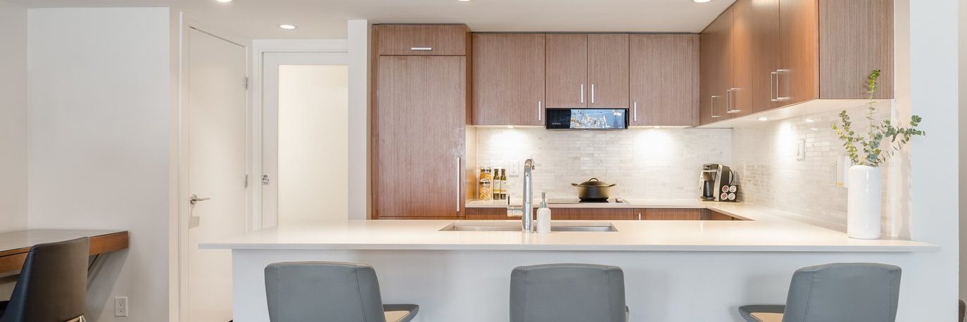 Modern kitchen with a white island countertop, three gray chairs, wooden cabinets, built-in appliances, and a vase with greenery on the right. The backsplash is white tile and the lighting is bright.