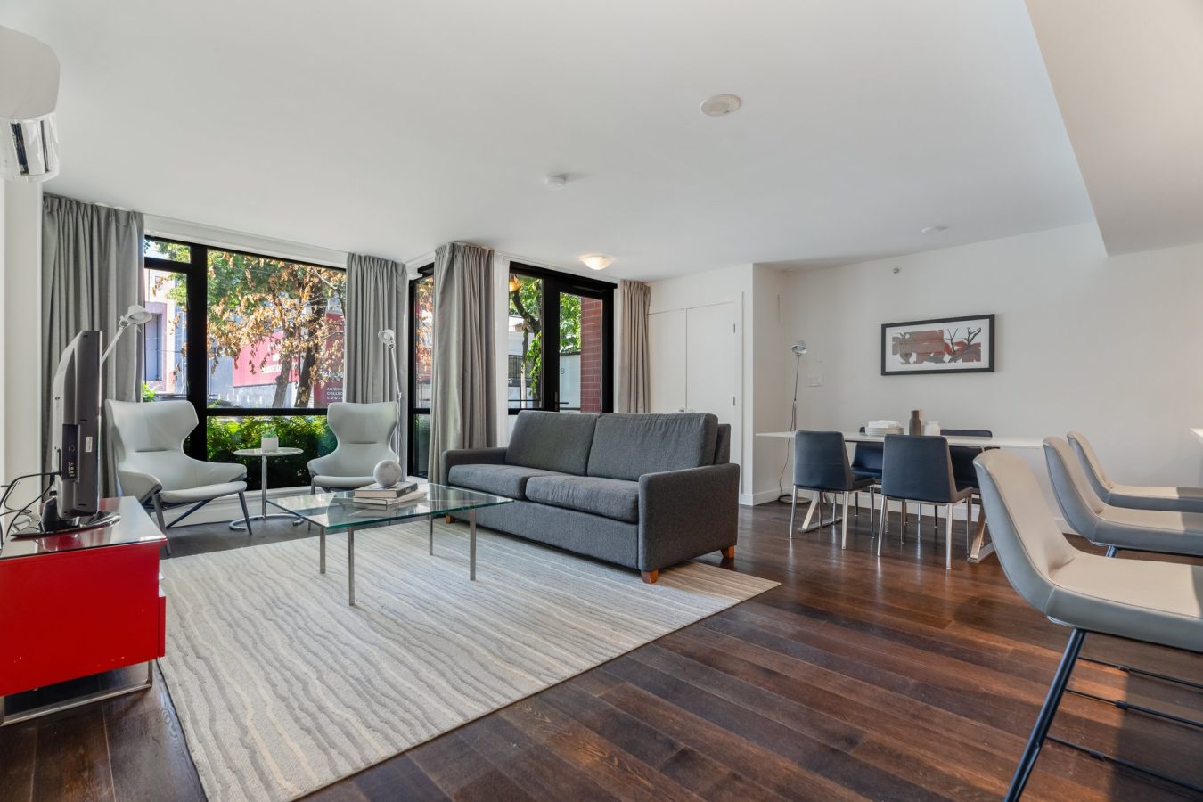 Modern living room with dark wood floors, a gray sofa, glass coffee table, two chairs by large windows, dining area with blue chairs, and a kitchen counter with white stools. Natural light fills the space.