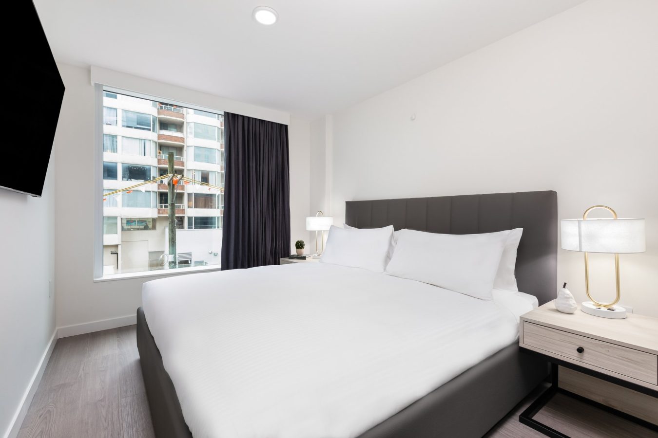 Modern hotel room with a large bed, white bedding, two bedside tables with lamps, and a window with city buildings and a construction crane visible outside. The room is bright with minimalistic decor.