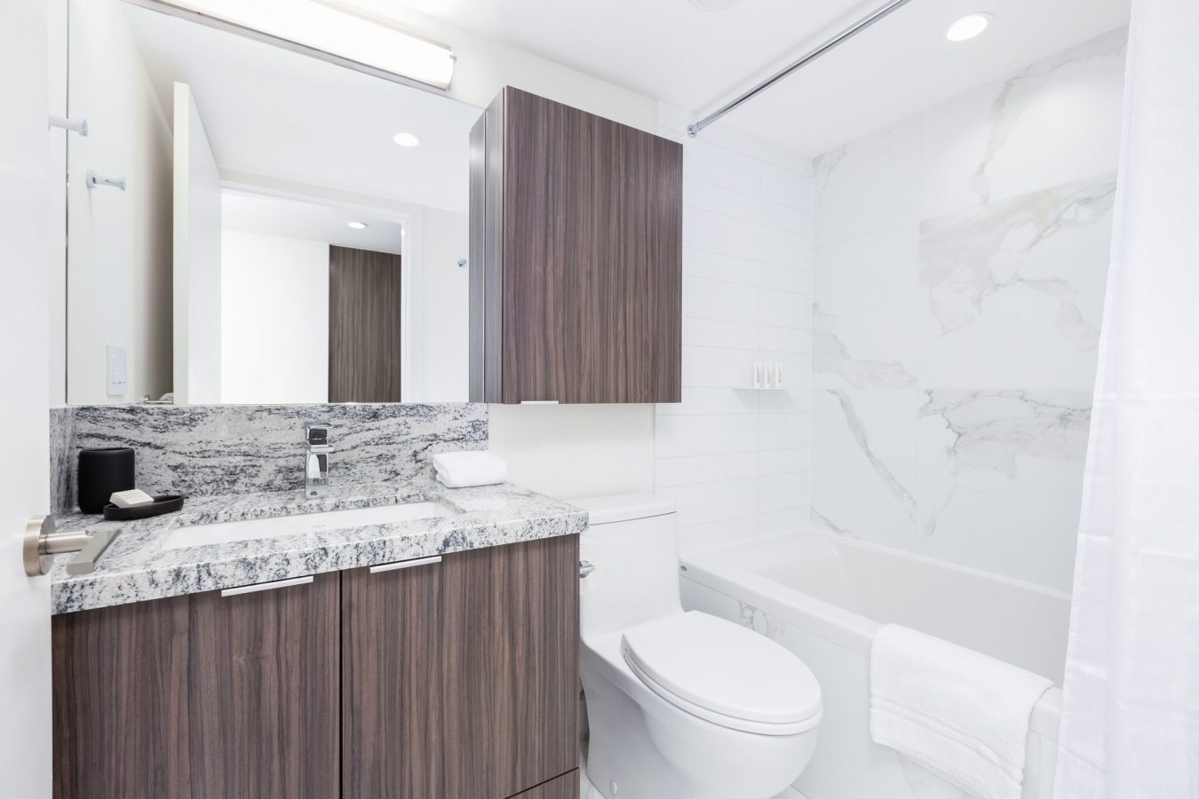 A modern bathroom with dark wood cabinets, a white toilet, a marble countertop, and a bathtub with marble-patterned tiles. A towel hangs on the bathtub, and the space is well-lit with overhead lights.