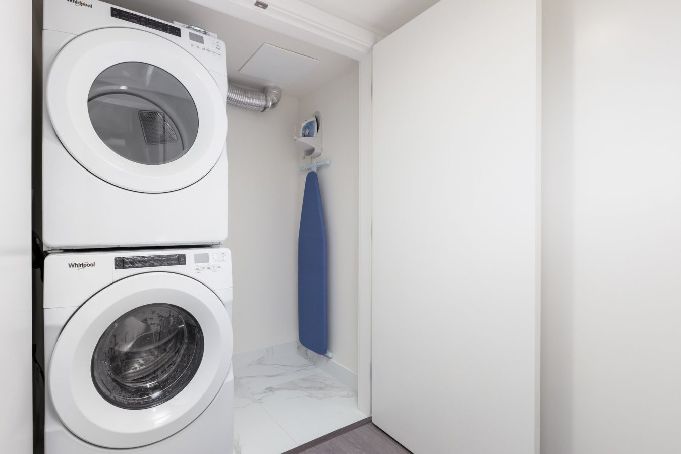 A laundry area with a stacked white washer and dryer next to a blue ironing board and iron, set against a light-colored wall and marble floor.