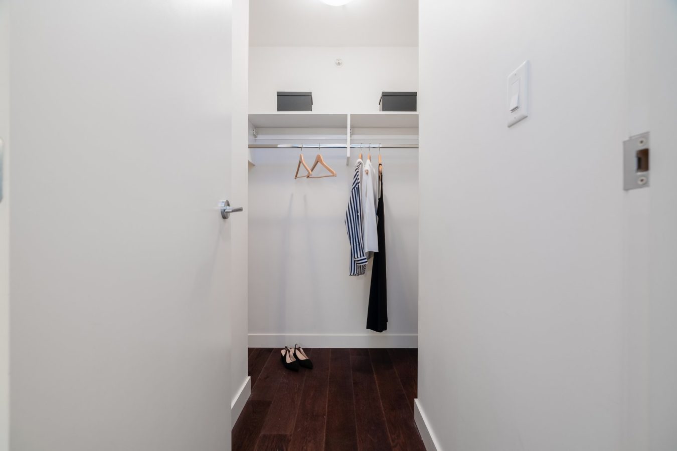 A walk-in closet with white walls, wooden floor, and a hanging rod. A few clothes and hangers are on the rod, a pair of shoes is on the floor, and two storage boxes are on the top shelf.