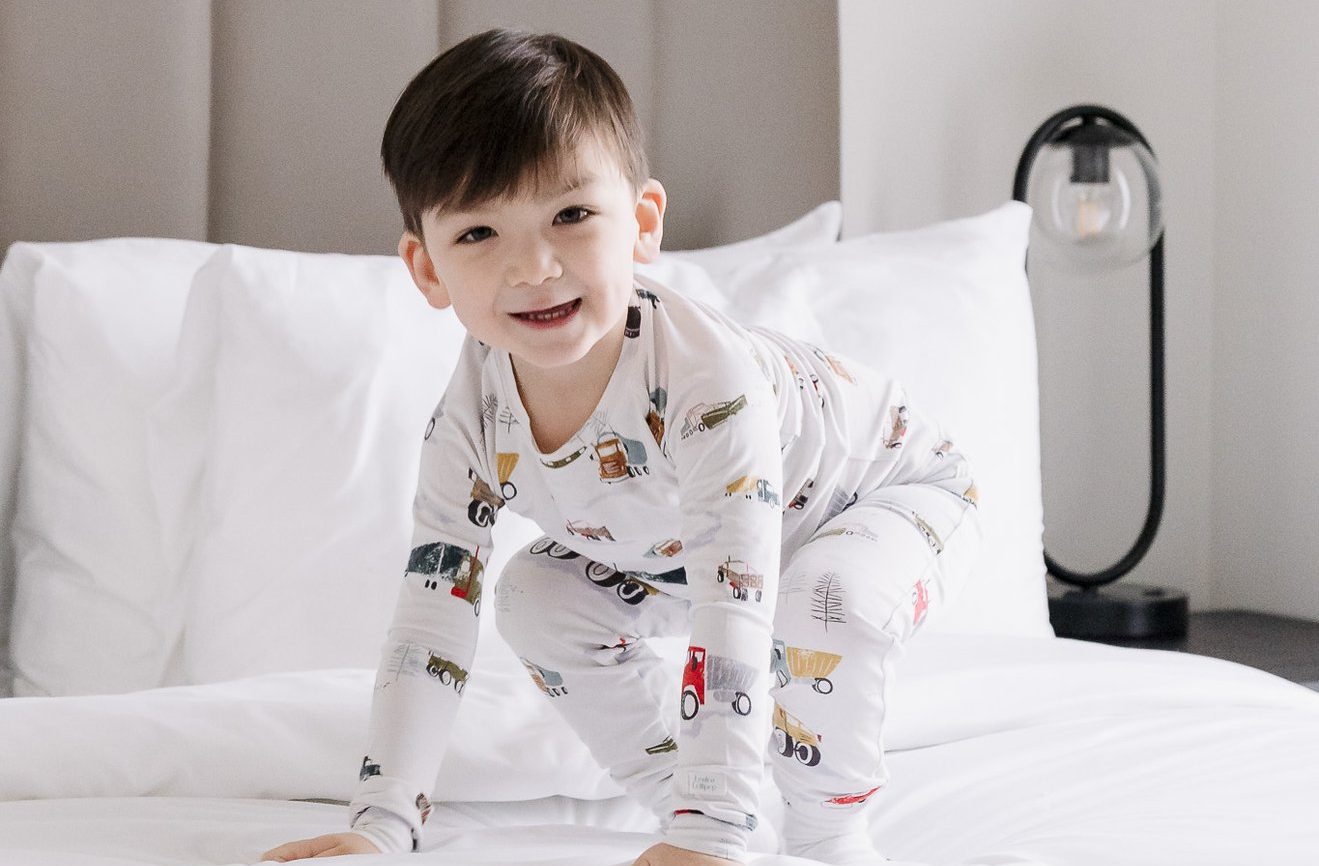 A young boy in patterned pajamas is smiling and kneeling on a white bed. A modern lamp is visible on a bedside table in the background.