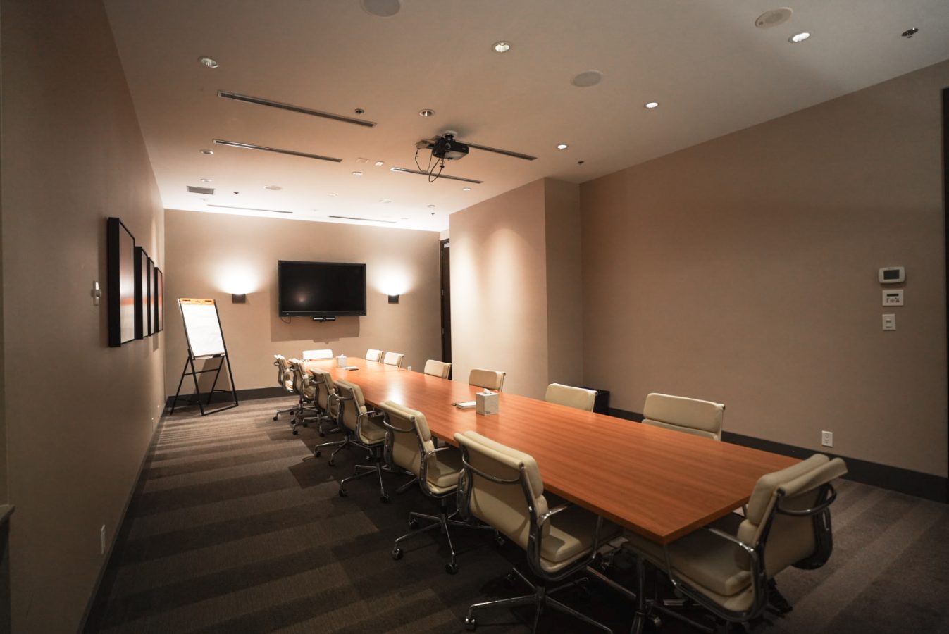 A modern conference room with a long wooden table, cream-colored chairs, a wall-mounted TV, a projector on the ceiling, and a flip chart at the end of the room. The walls are neutral-colored and the lighting is bright.