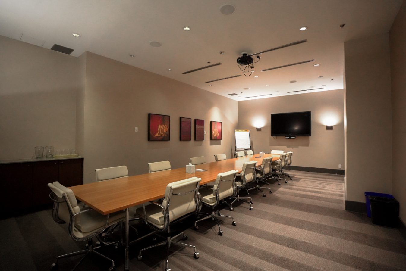 A modern conference room with a long wooden table, white office chairs, a wall-mounted TV, a projector on the ceiling, framed artwork on the wall, and a flip chart at the far end.