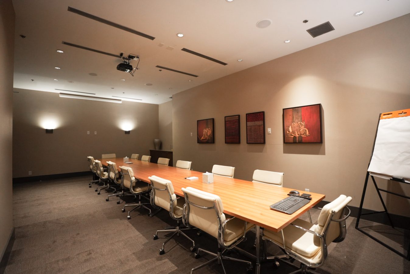 A modern conference room with a long wooden table, white office chairs, wall art, a projector on the ceiling, and a flip chart stand. The room is softly lit with recessed and wall lights.
