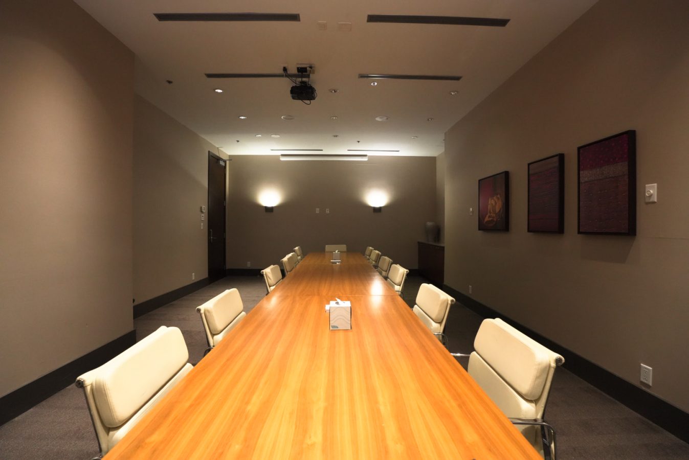 A modern conference room with a long wooden table surrounded by white chairs, beige walls, ceiling lights, wall art on the right, and a projector mounted above the table.