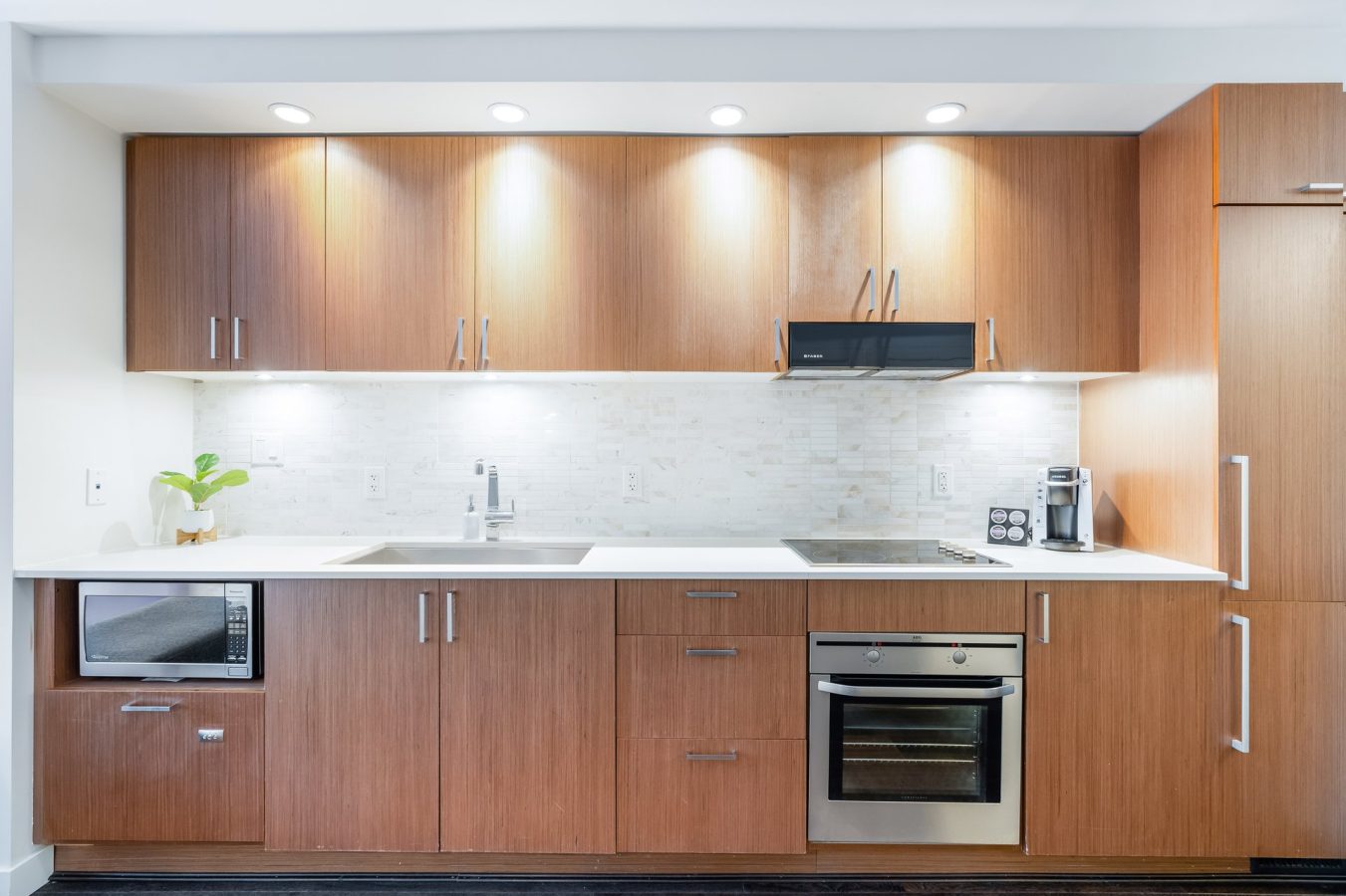 Modern kitchen with wood cabinets, white countertop, built-in oven, stovetop, microwave, sink, coffee maker, and a small green plant. White tile backsplash and under-cabinet lighting create a bright, clean look.
