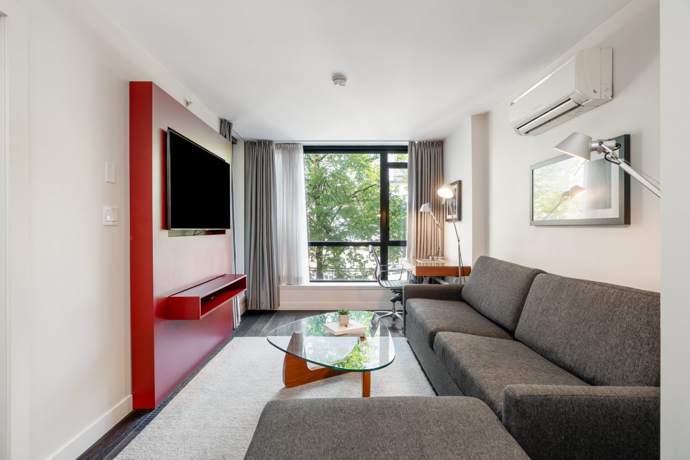 Modern living room with gray sofas, a glass coffee table, a wall-mounted TV on a red accent wall, large window with curtains, wooden desk, chair, and lamps. Tree foliage is visible outside through the window.