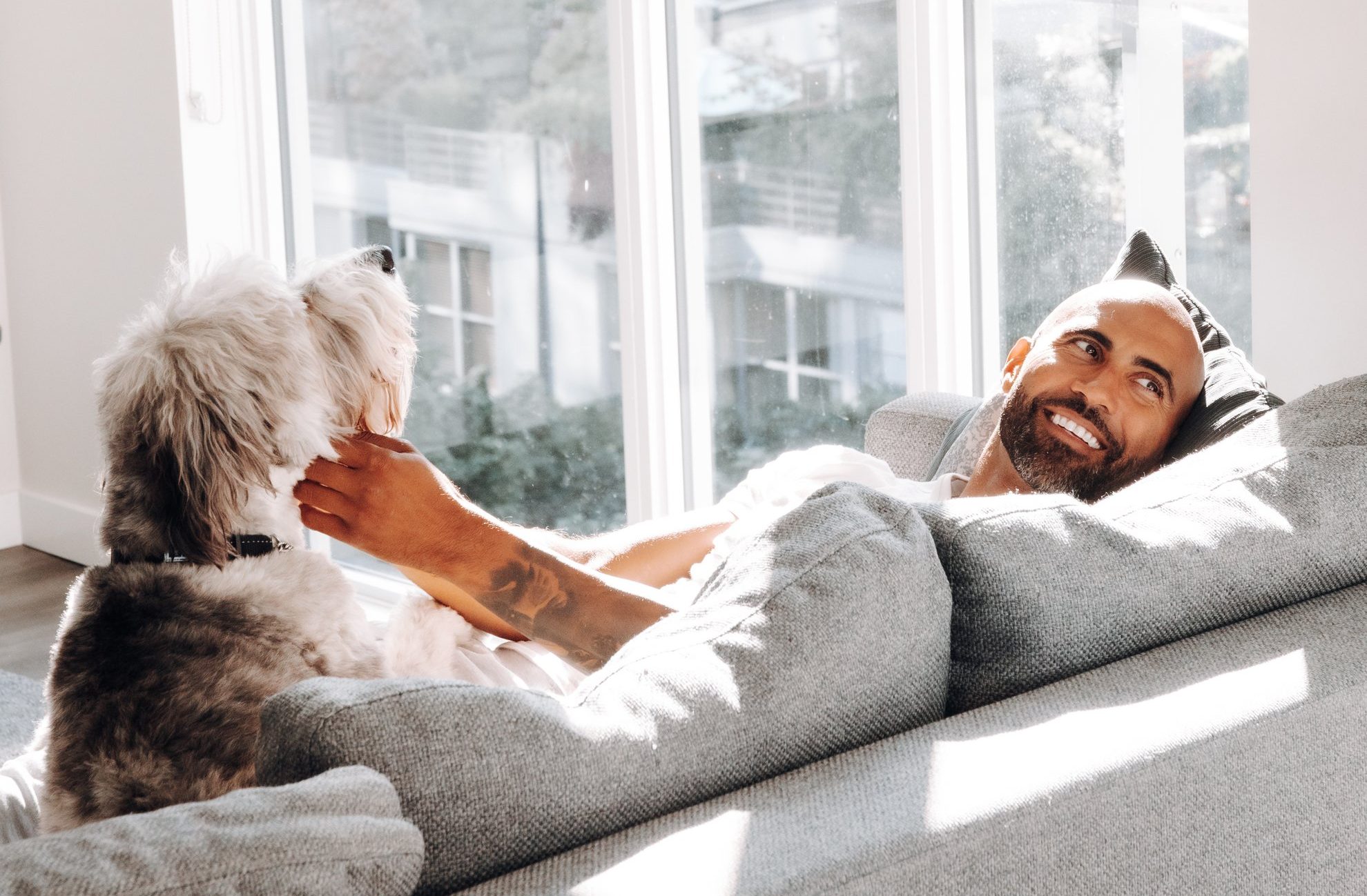 A man relaxes on a grey couch, smiling and gently petting a fluffy dog by his side. Sunlight streams through large windows behind them, creating a warm, cheerful atmosphere.