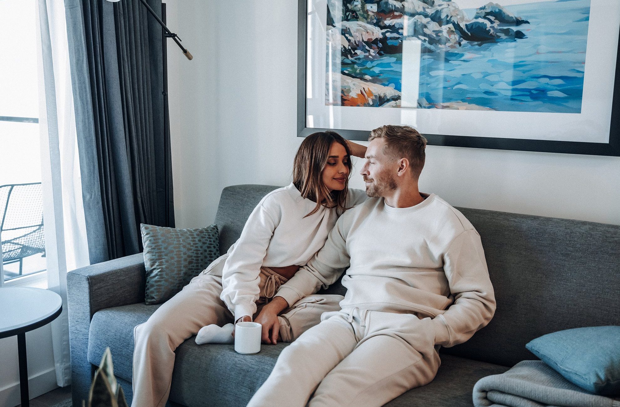 A couple sits closely on a gray sofa in a modern living room, wearing matching light-colored loungewear. The woman holds a white mug, and they smile at each other. A painting of water and rocks hangs on the wall behind them.