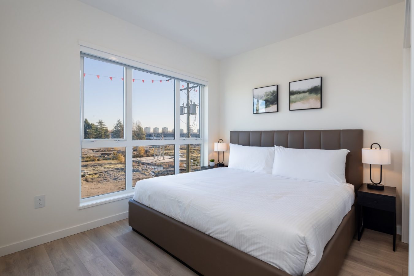 A bright, modern bedroom with a large bed, white bedding, two nightstands with lamps, and two framed pictures above the headboard. Sunlight streams through a large window with a view of trees and buildings outside.