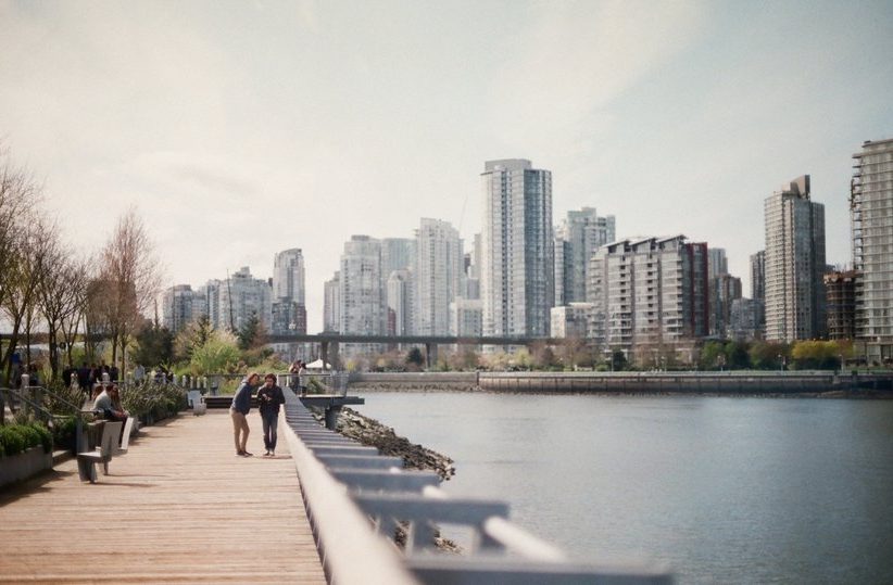 People walk along a wooden boardwalk beside a body of water, with a city skyline of modern high-rise buildings in the background under a lightly cloudy sky. Trees and benches line the pathway.