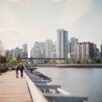 Two people walk together on a wooden boardwalk next to a calm body of water, with a cityscape of modern high-rise buildings in the background under a partly cloudy sky.