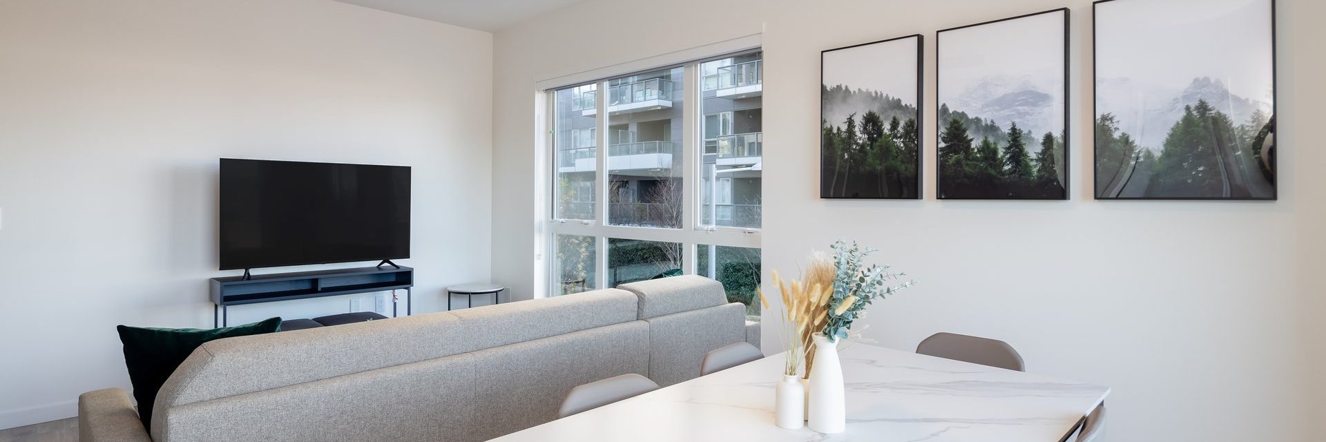 A modern living room with a gray sofa, flat-screen TV, large window, and three forest landscape art pieces on the wall, next to a dining table with vases and dried flowers.