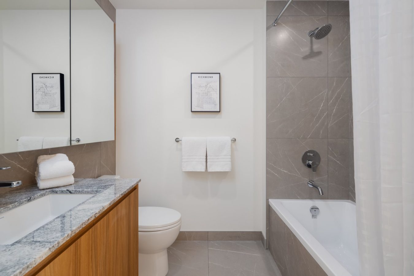 Modern bathroom with gray tile, a bathtub with shower, white curtain, toilet, wood vanity with a granite countertop, mirror cabinet, towels, and framed artwork on the white wall.