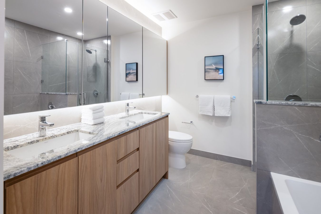Modern bathroom with gray tile, double sink vanity with marble countertop, large mirrors, glass shower, bathtub, toilet, two framed pictures, and folded white towels on counter and rack. Bright, clean, and minimalist design.