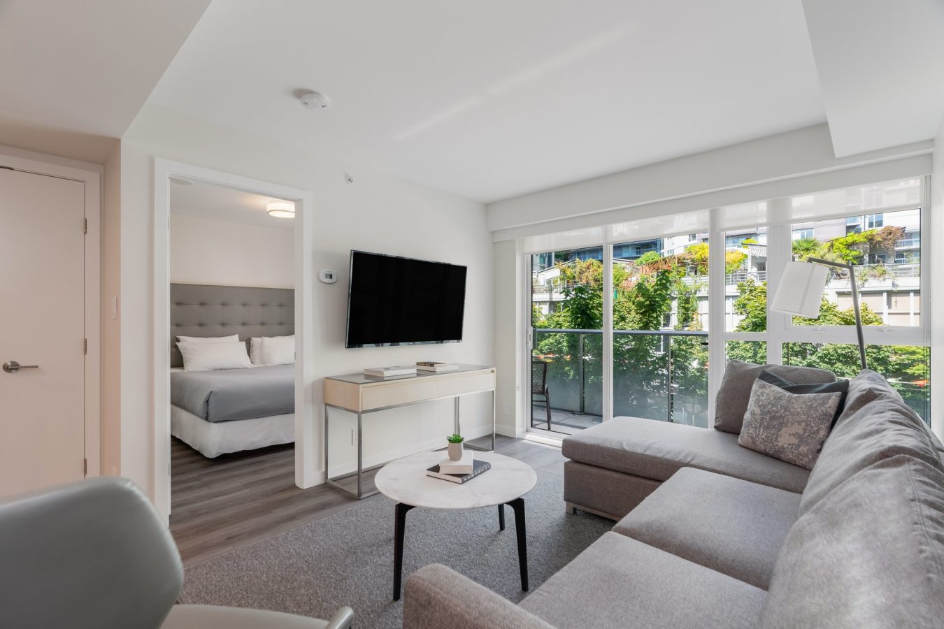 A modern, bright living room with a gray sectional sofa, round coffee table, wall-mounted TV, and large windows, with a view into a bedroom featuring a neatly made bed and tufted headboard.