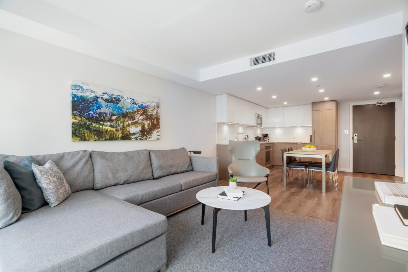 Modern living room with a gray sectional sofa, round coffee table, abstract wall art, and open kitchen in the background with white cabinets, wood flooring, and a dining area.