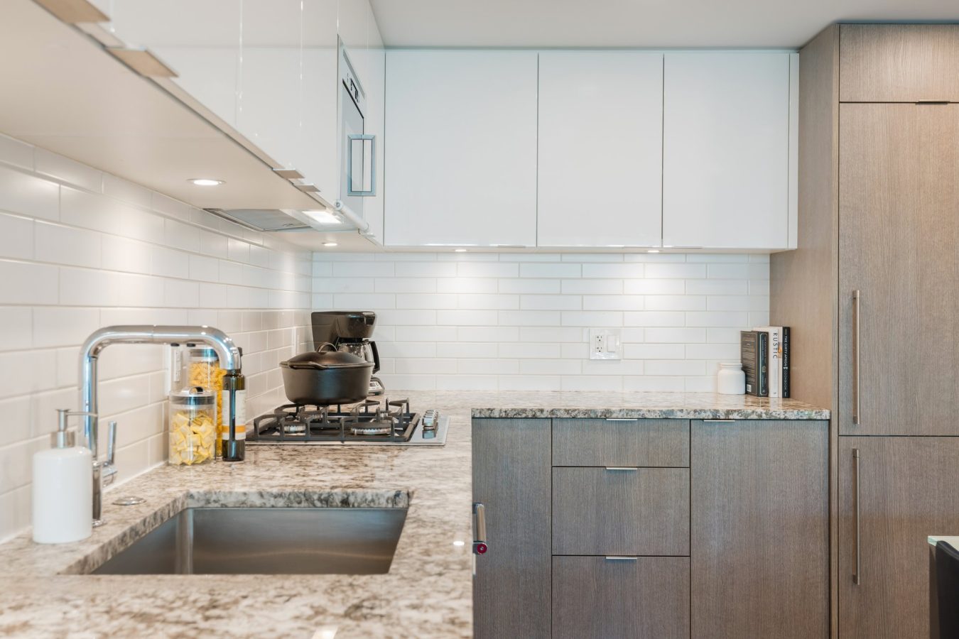 Modern kitchen with granite countertops, a stainless steel sink, stovetop with pots, white subway tile backsplash, light wood cabinets, and white upper cabinets. A jar of pasta and kitchen essentials are on the counter.