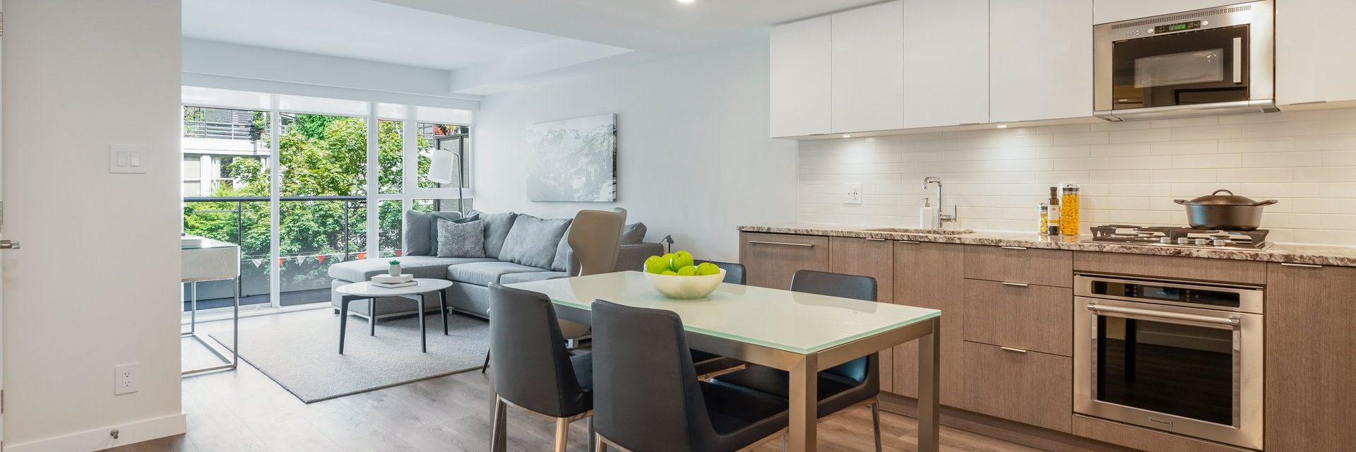 Modern apartment with an open-plan kitchen, dining area, and living room. Glass dining table with black chairs, gray sofa, and large window letting in natural light. White cabinets and stainless steel appliances in the kitchen.