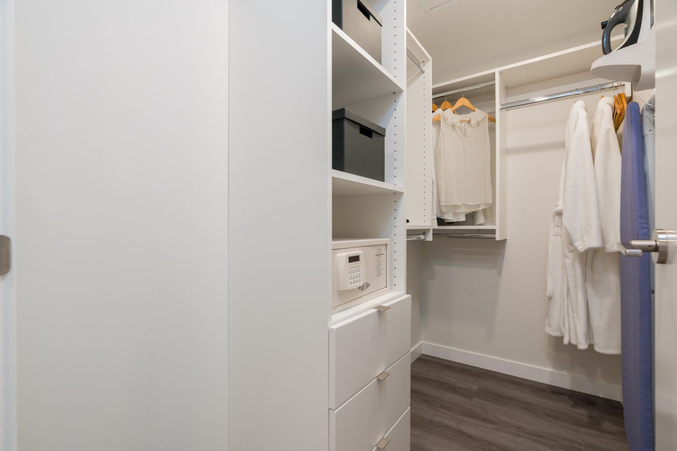 A modern walk-in closet with white shelves, drawers, storage boxes, a safe, hanging white shirts on wooden hangers, and white robes. The space has light walls and dark wood flooring.