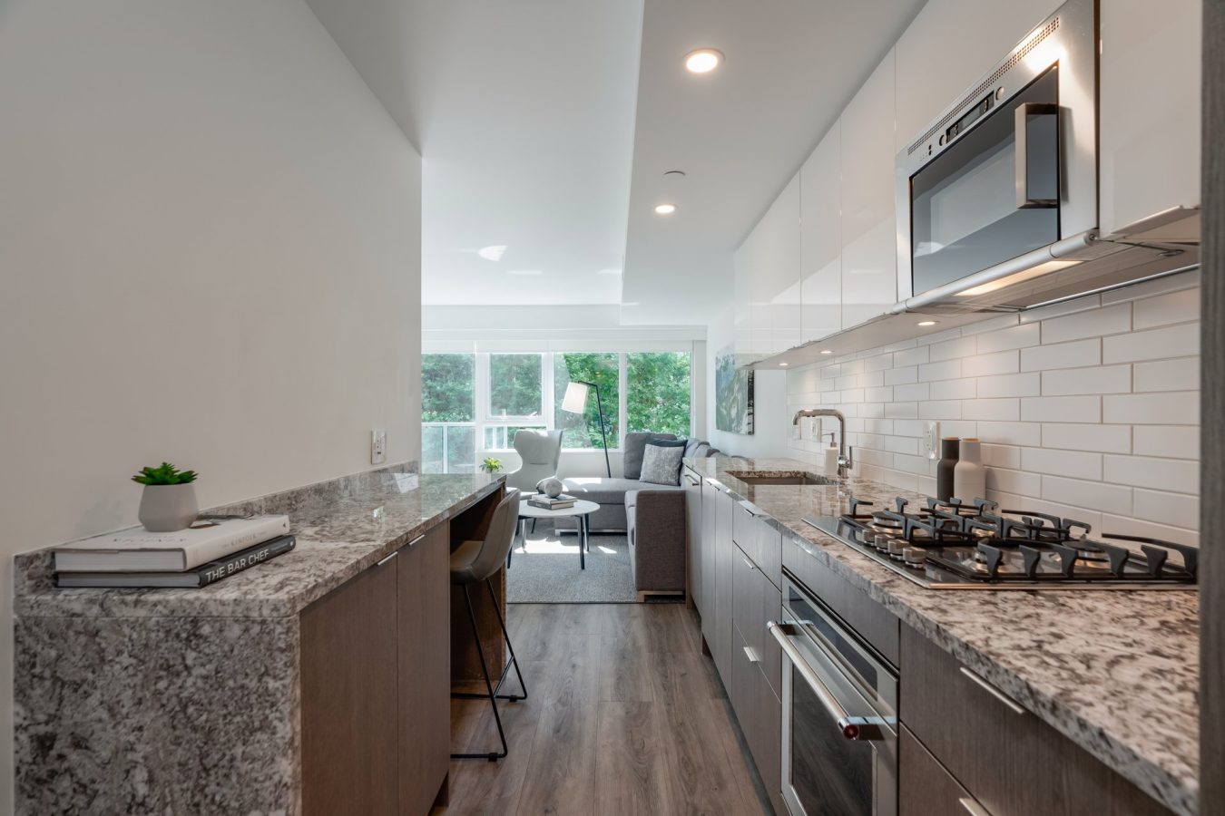 Modern galley kitchen with granite countertops, gas stove, white cabinets, and subway tile backsplash, opening into a bright living area with large windows and a green view outside.