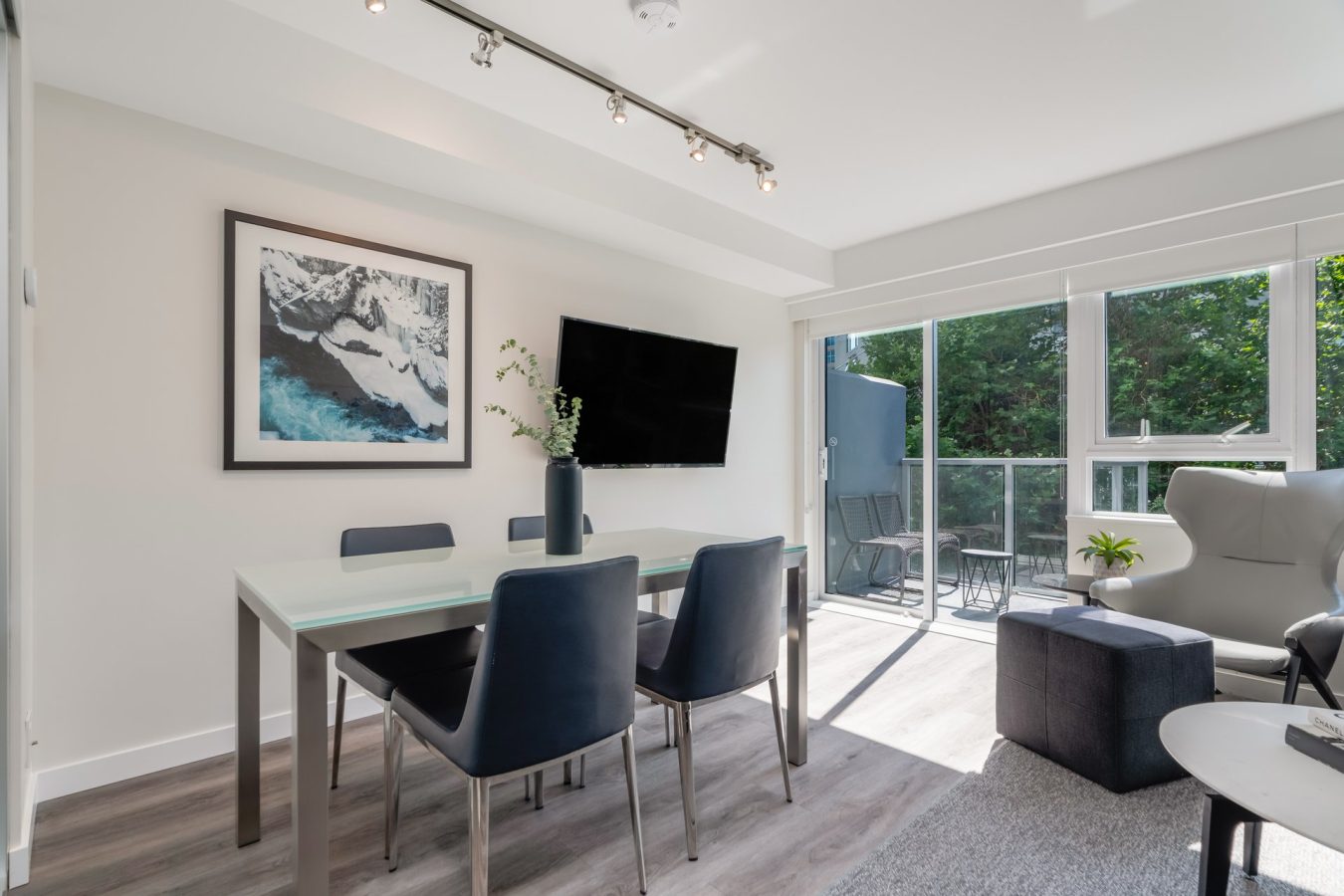 Modern dining and living area with a glass table, four navy chairs, wall art, mounted TV, large windows, and a balcony with outdoor seating; natural light fills the space.