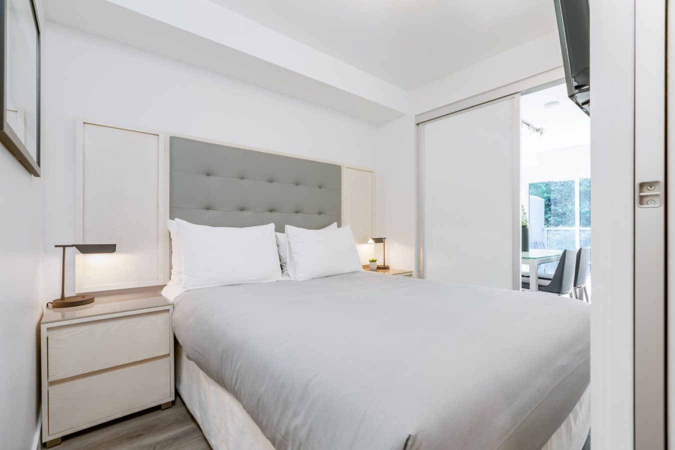 Modern, minimalistic bedroom with a bed featuring a grey tufted headboard, white bedding, two bedside tables with lamps, light wood flooring, and a sliding door opening to a bright adjoining room.