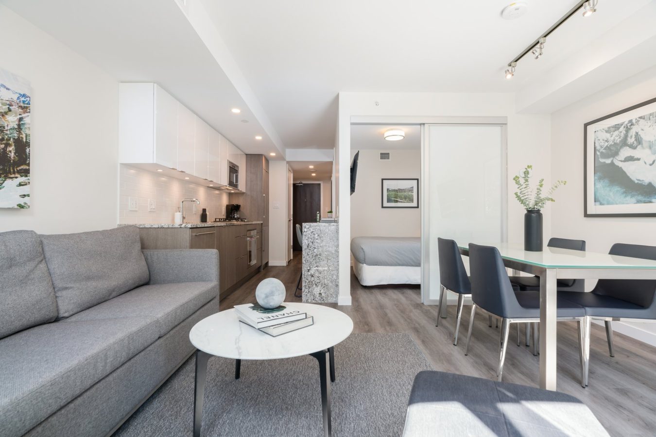 Modern studio apartment with a gray sofa, coffee table, open kitchen, and dining area. A bed is visible through sliding glass doors. Light wood floors and large windows create a bright, airy space with minimalist decor.