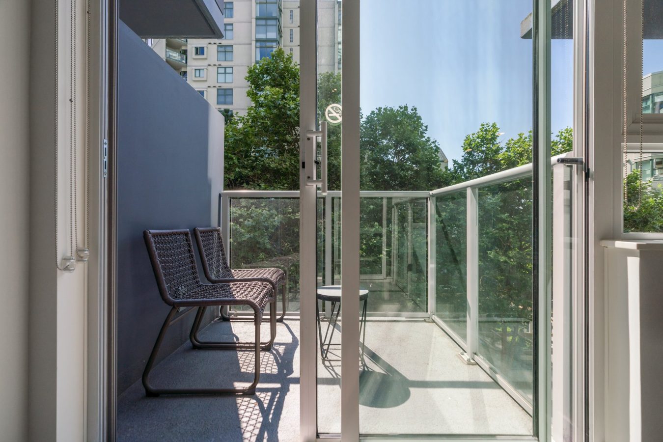 A modern balcony with glass railings, two wicker chairs, and a small table, seen through an open sliding door. High-rise buildings and green trees are visible in the background on a sunny day.