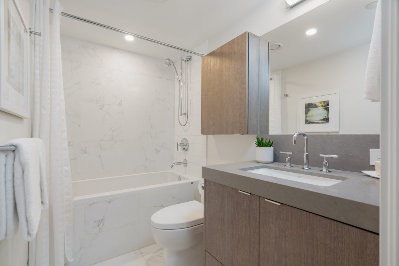 Modern bathroom with light wood vanity, white countertop, built-in sink, large mirror, wall-mounted cabinet, toilet, and a bathtub with marble tile surround. A plant and a towel add decorative touches.