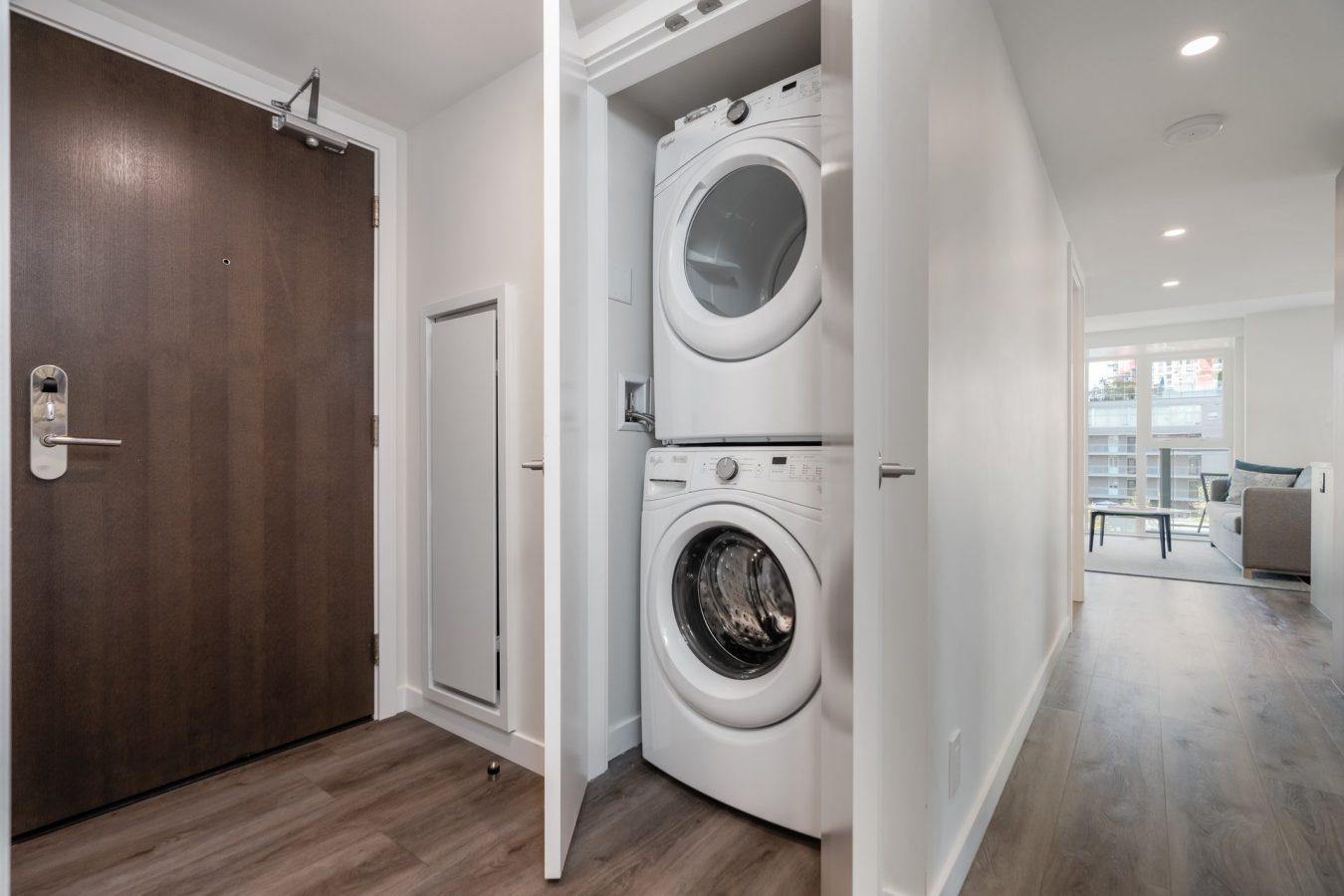 A stacked washer and dryer are tucked inside a closet with white doors in a modern hallway with wood flooring, near a dark wooden entrance door and a living area with large windows.