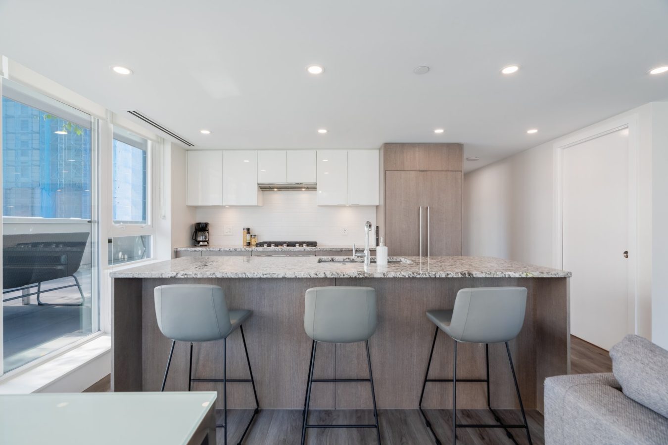 Modern kitchen with a large island featuring a granite countertop, three gray barstools, white cabinets, built-in appliances, and large windows letting in natural light.