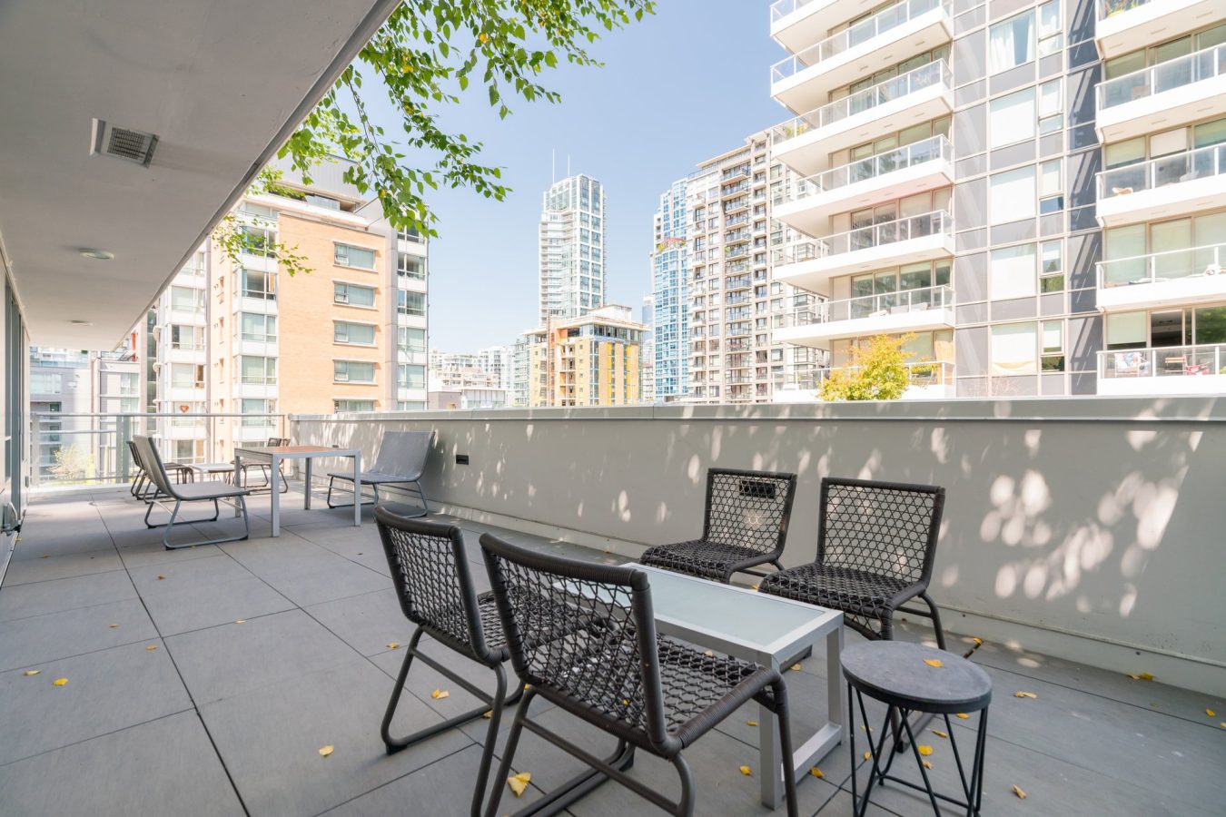 A spacious urban balcony with modern black chairs and a white table, surrounded by tall apartment buildings on a sunny day. Some leaves are scattered on the tiled floor and green tree branches overhang above.